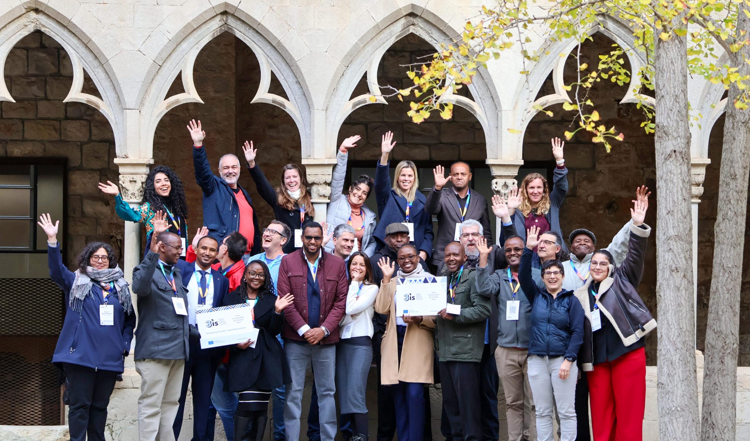 A large group of 3is project partners stands together in an outdoor courtyard, smiling and raising their hands in celebration.