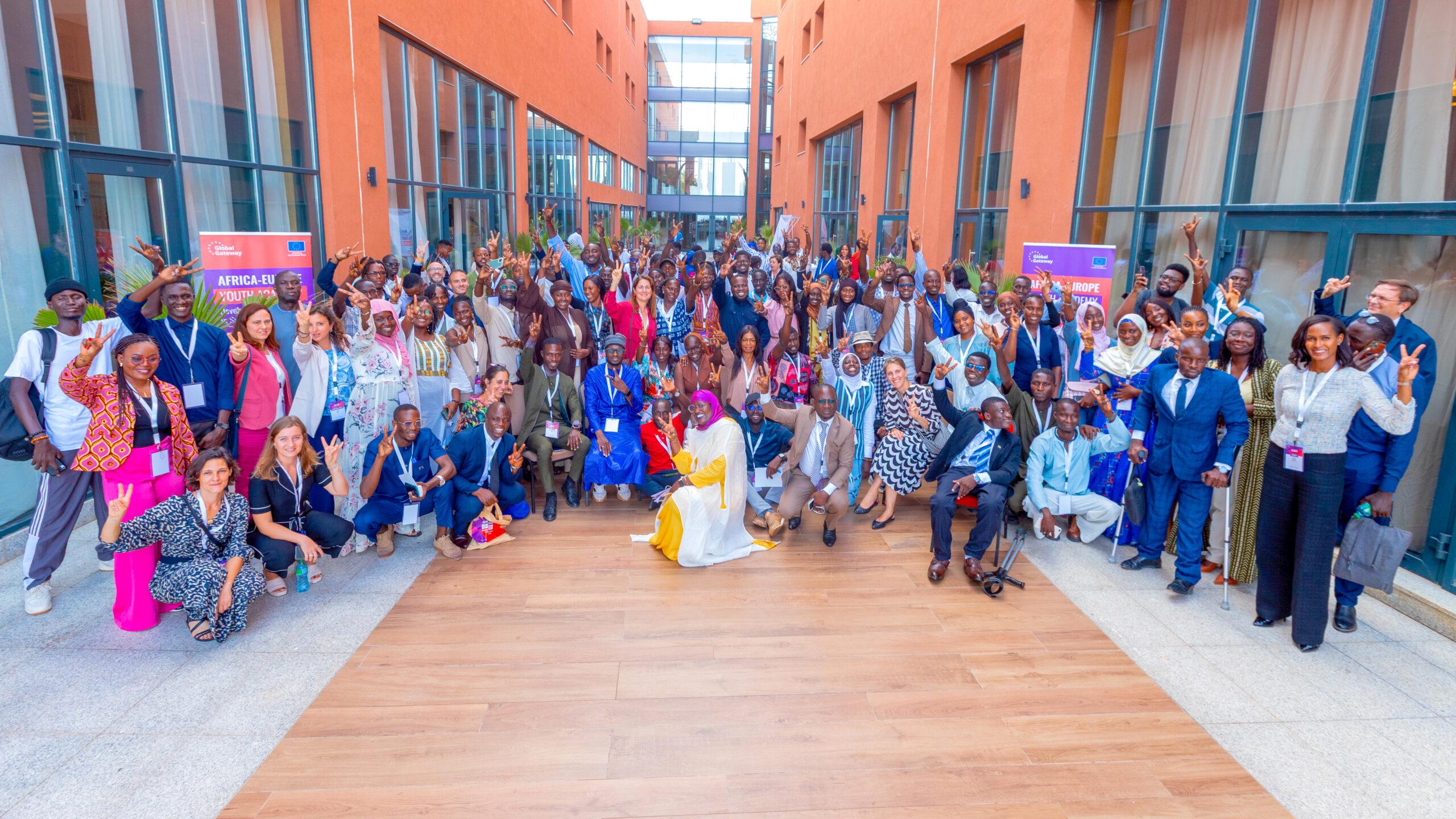 Large group of diverse participants at an Africa-Europe youth event posing together outdoors, smiling and making peace signs between modern orange buildings.
