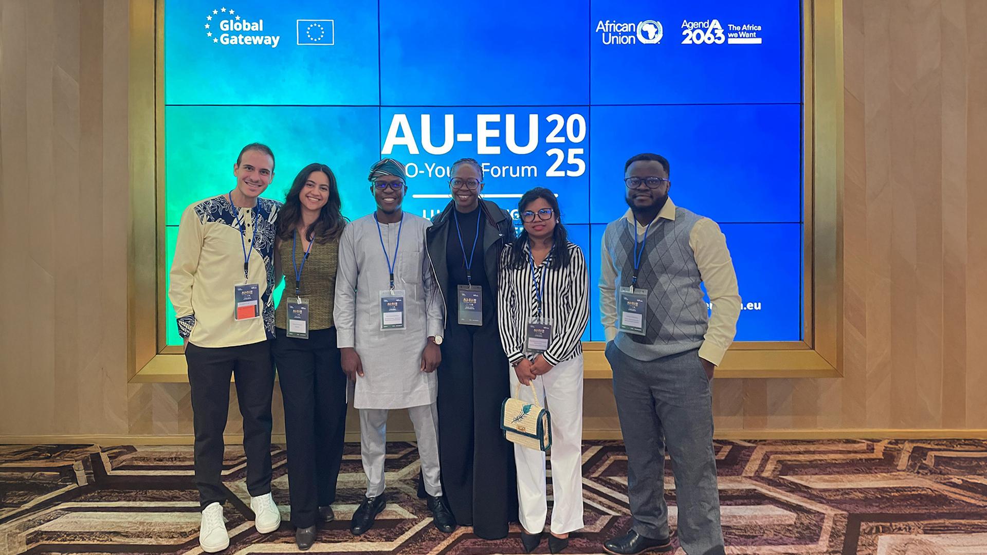 Six participants stand together indoors in front of a large digital display wall featuring the branding of the AU–EU Youth Forum 2025. The backdrop shows the Global Gateway logo, the African Union emblem, and references to Agenda 2063.