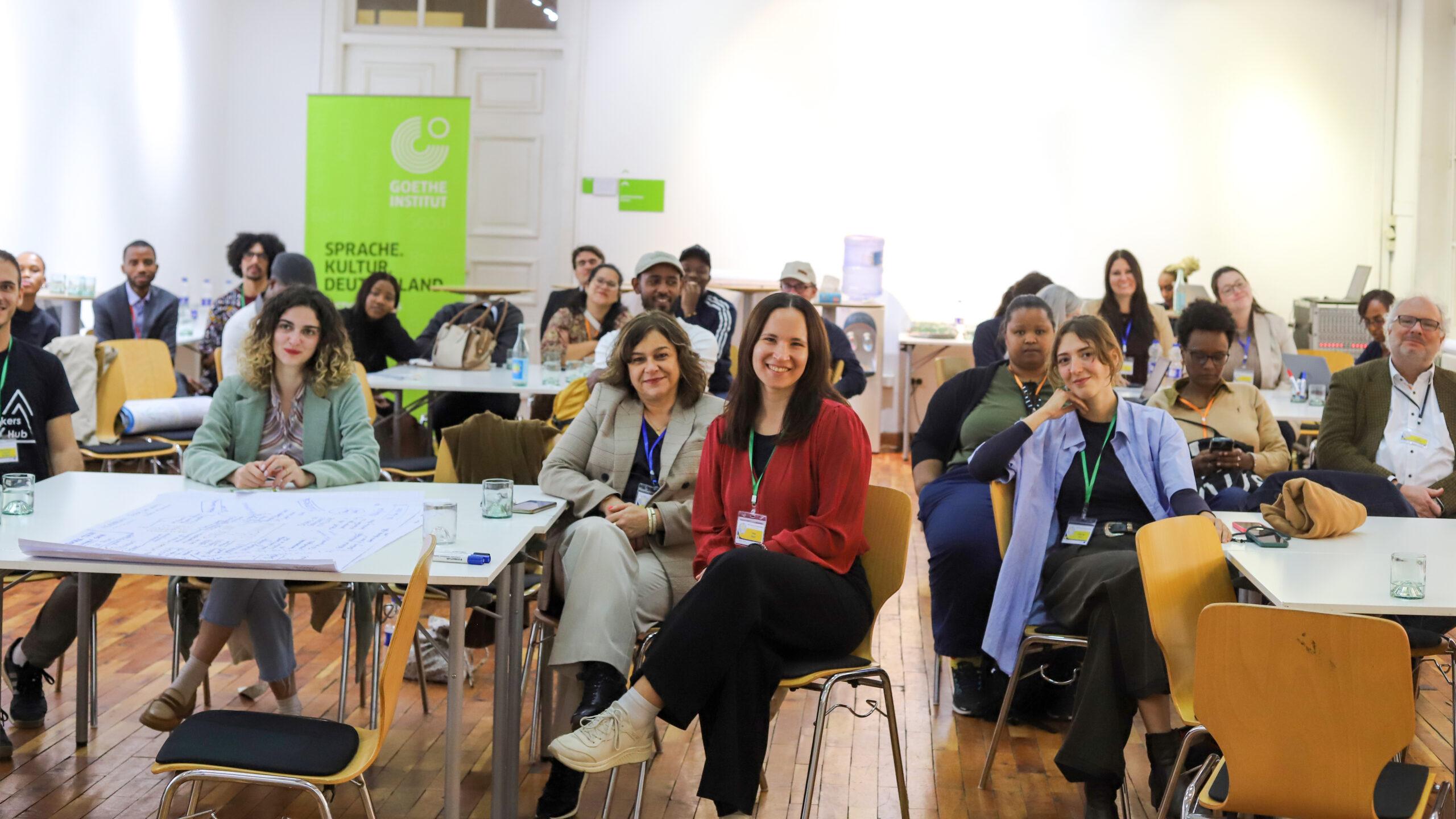Group of diverse participants seated in a workshop setting, smiling and facing the camera, with a Goethe-Institut banner in the background.