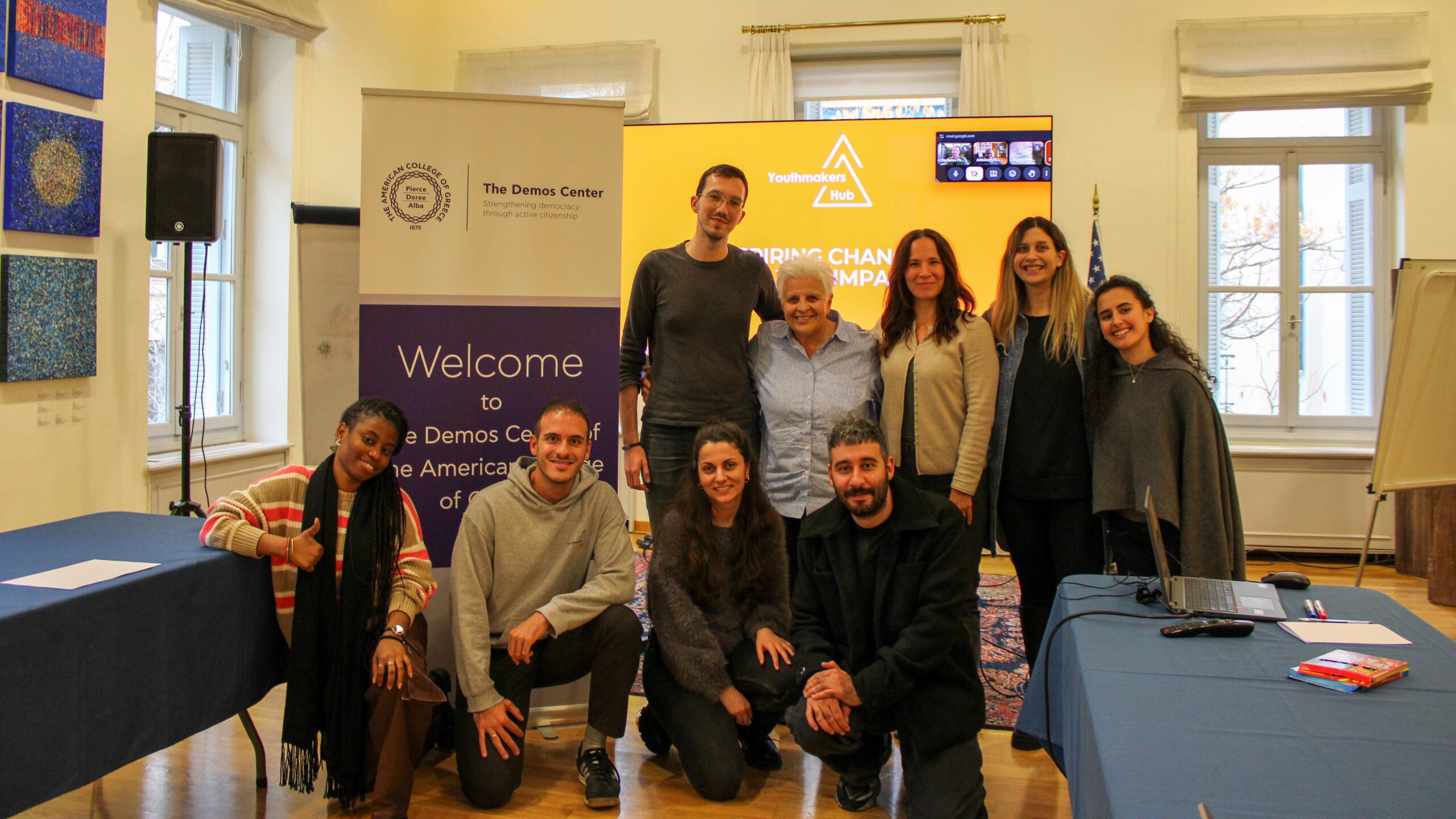 Youthmakers Hub team posing together at the Demos Center during a meeting, with presentation screen and welcome banner in the background.