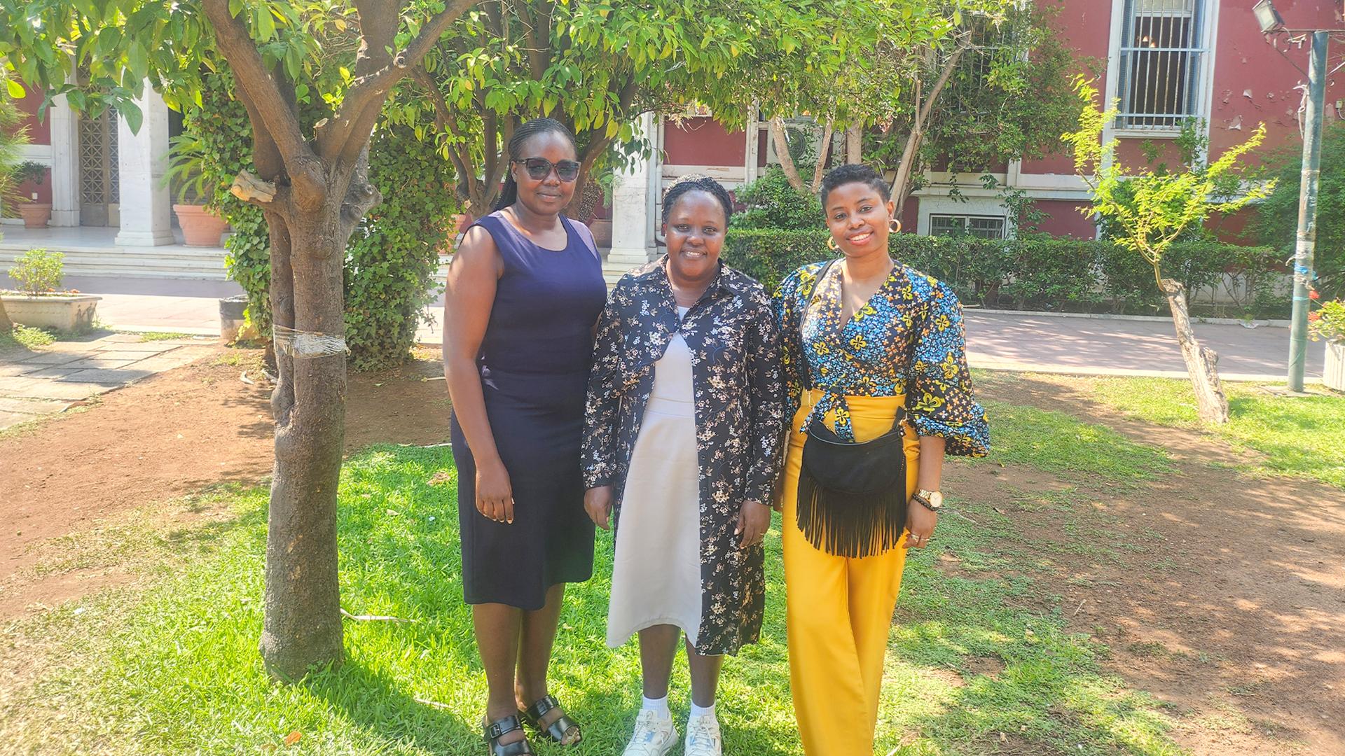 Three women stand together outdoors on grass near trees and a red building, smiling at the camera on a sunny day.
