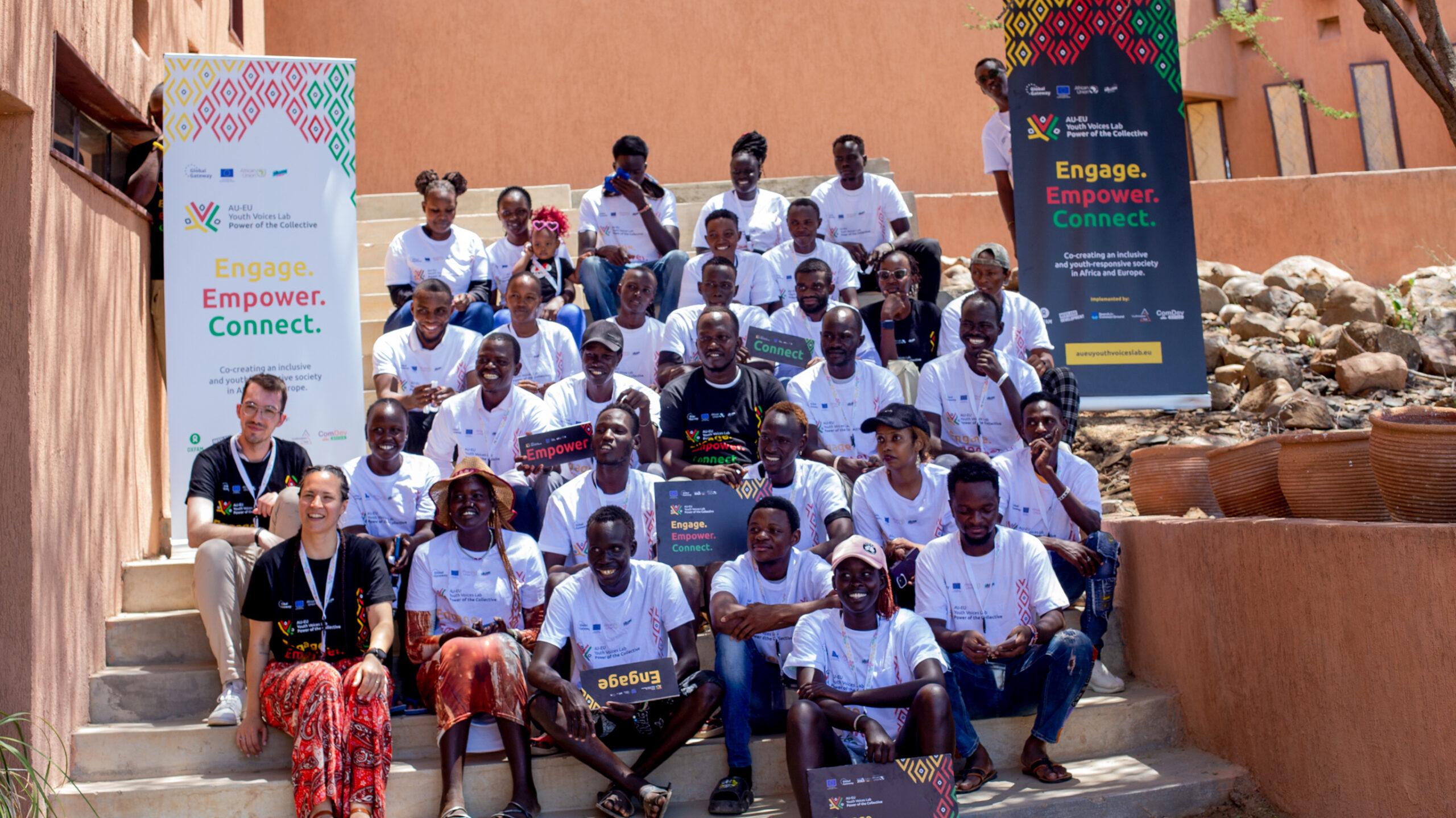 A large group of young people sit together on outdoor steps in front of a building, posing for a group photo. Banners stand on each side of the group, displaying the branding of the “AU–EU Youth Voices Lab” with colorful geometric patterns and the tagline “Power of the Collective – Engage. Empower. Connect.