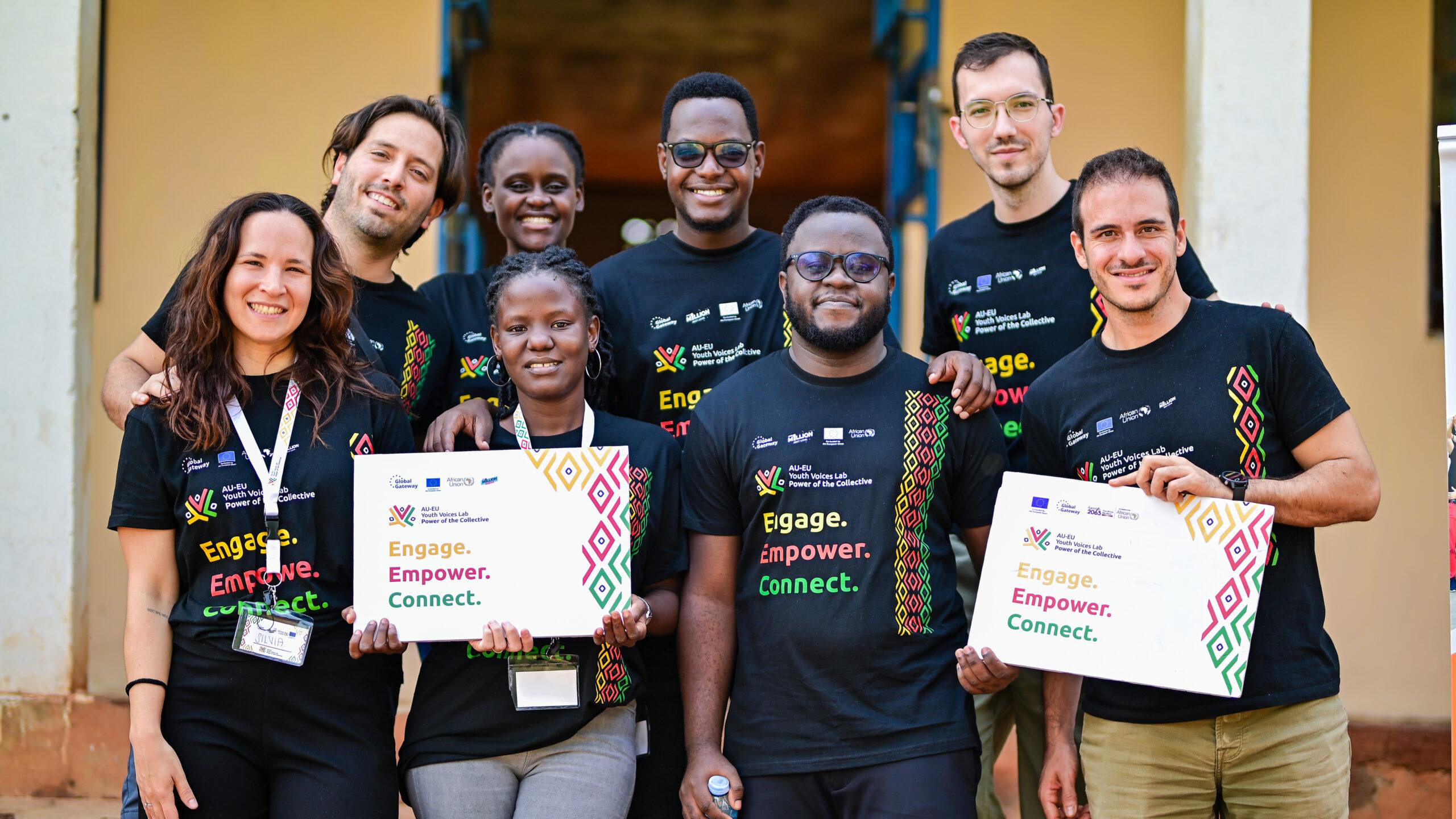 Team of 8 trainers posing for a group photo during the “Lead the Narrative; Communication for Youth Advocacy Training in Uganda. They are wearing AU-EU Youth Voices Lab - Power of the Collective shirts and are holding props.