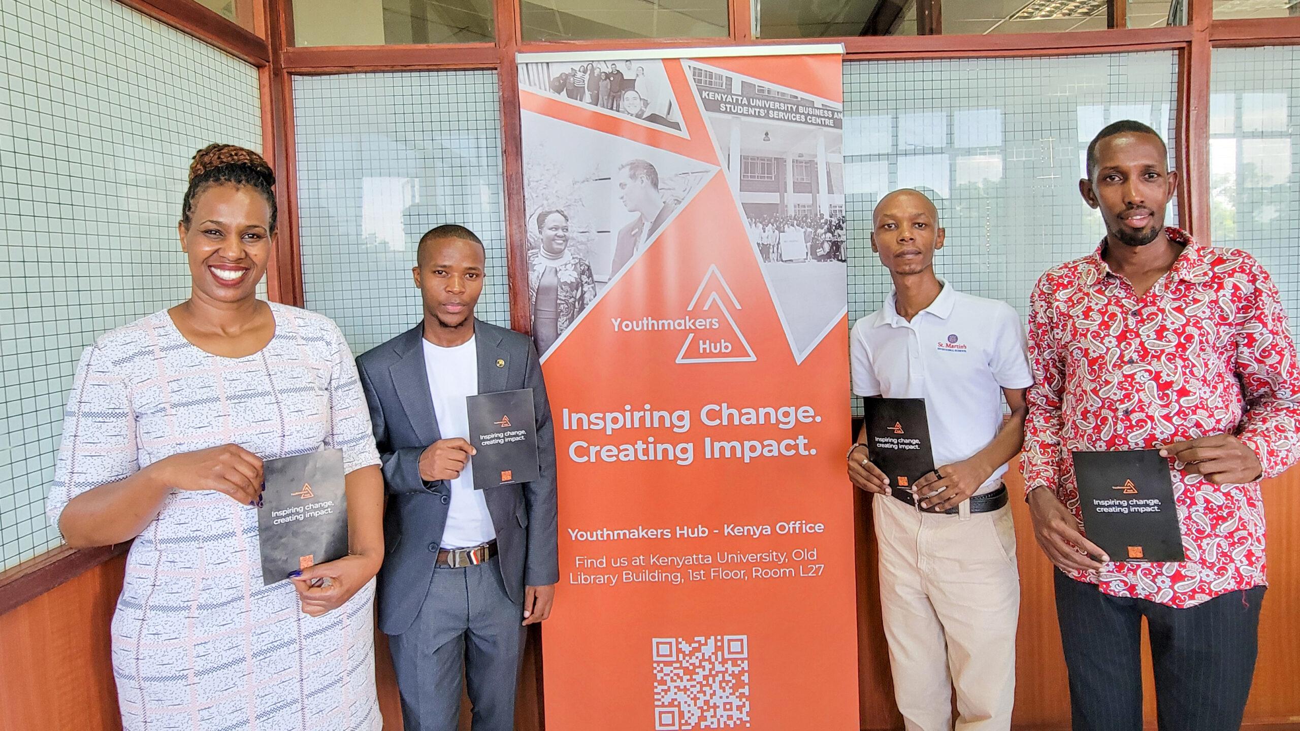 Four young professionals stand indoors holding black Youthmakers Hub booklets. Behind them is an orange-and-white banner announcing the Youthmakers Hub Kenya Office, located at Kenyatta University’s Building.